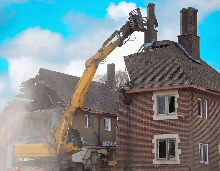 Excavator tearing down a brick house during demolition project. Dust and debris visible.