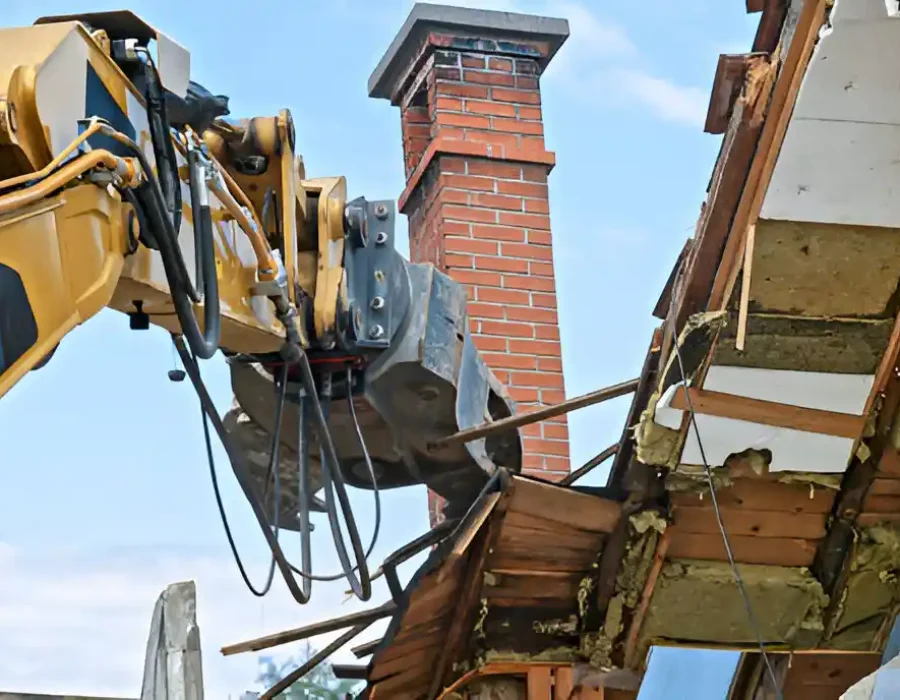 Excavator tearing down a house roof and chimney during demolition.