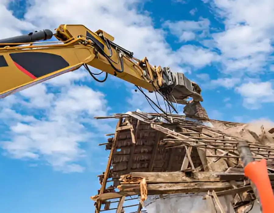 Excavator tearing down an old wooden building under a clear blue sky.