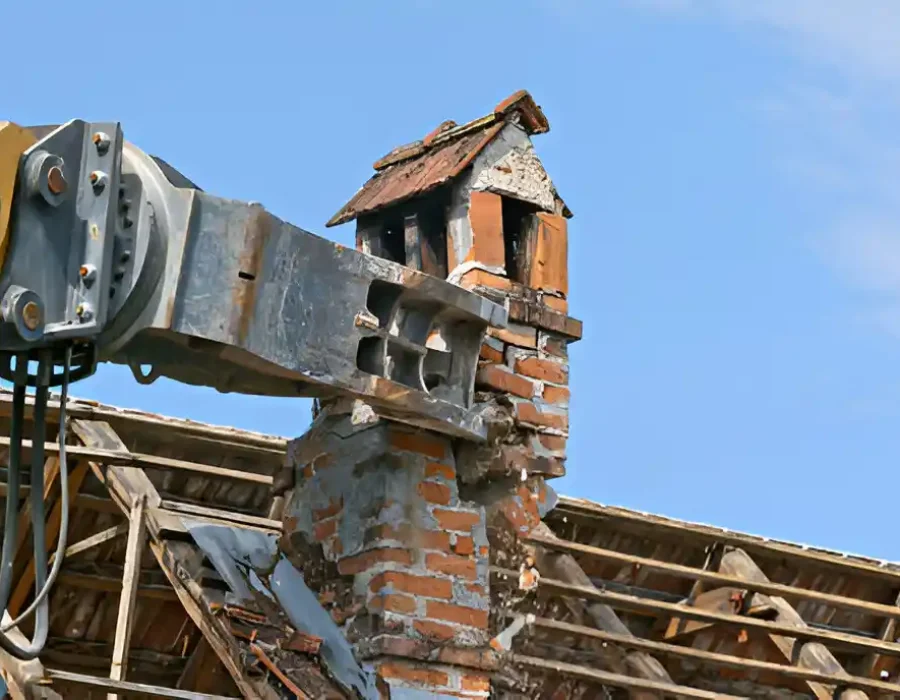 Excavator arm demolishing a brick chimney on a damaged roof under clear blue skies.