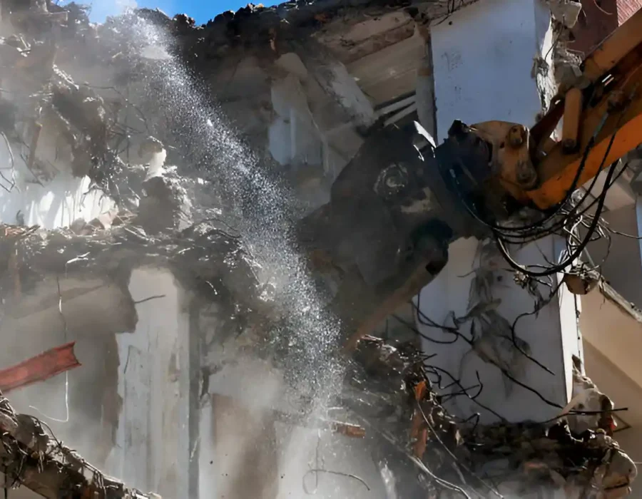 Excavator demolishing a building with water spray for dust suppression.