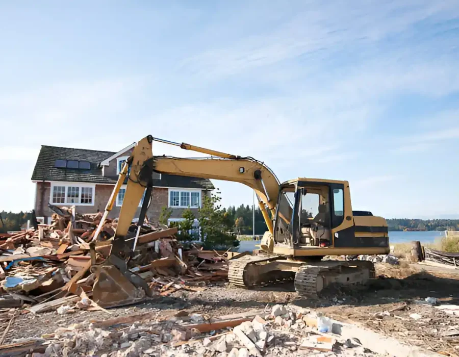 Excavator removing debris at a residential demolition site near a house and waterfront.
