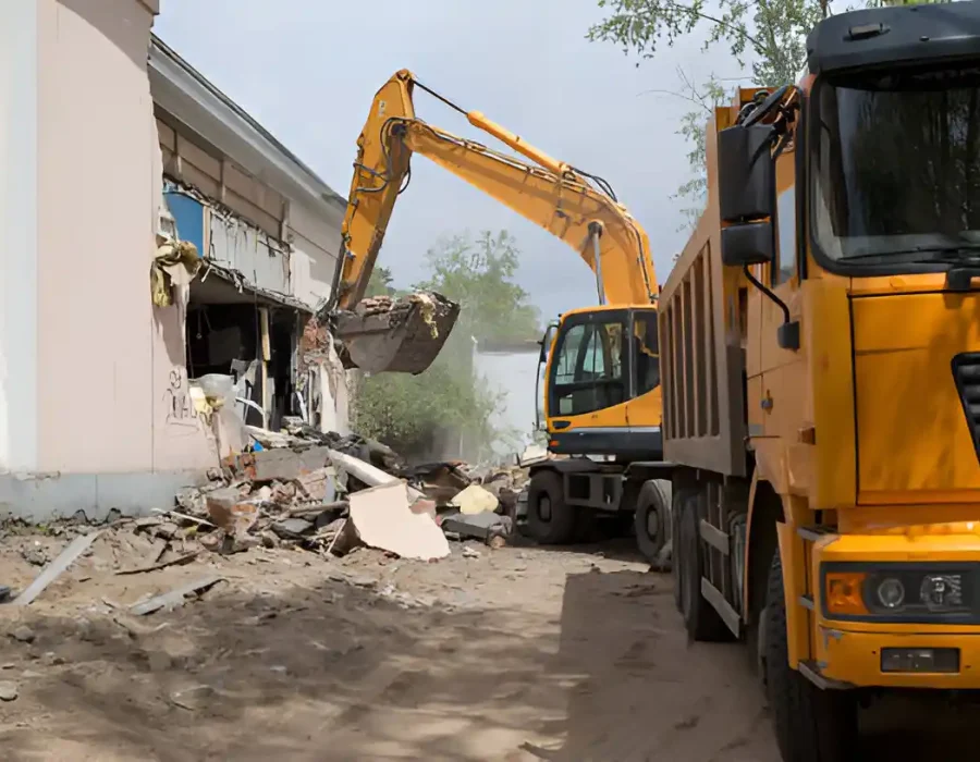 Excavator tearing down a building with a truck for debris removal nearby.