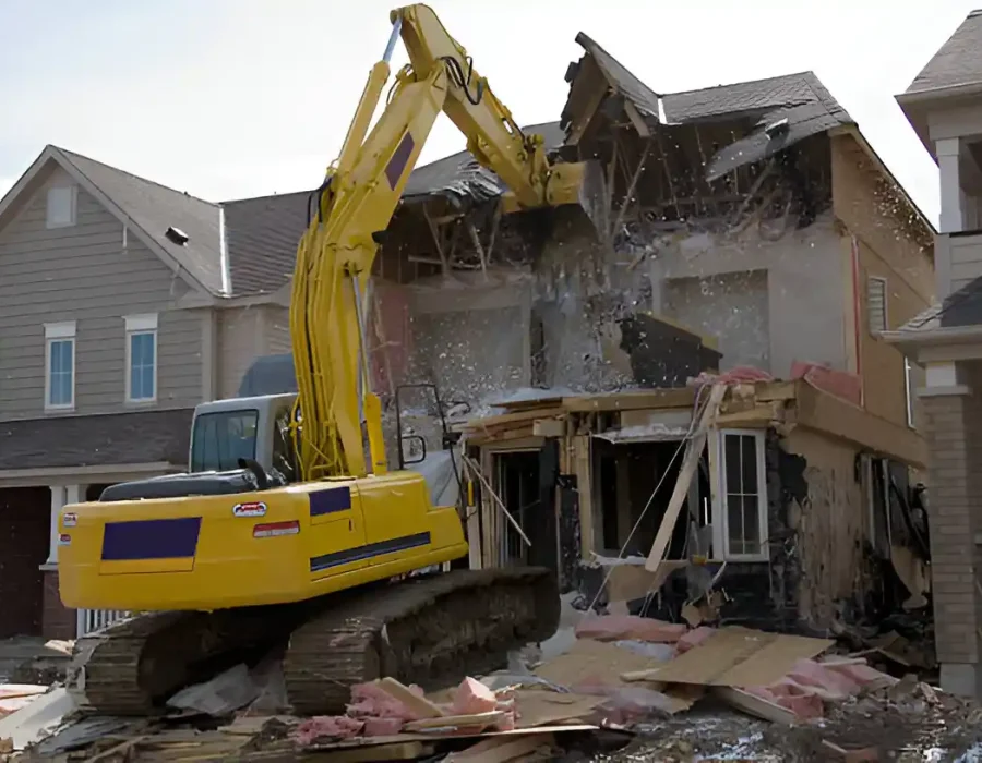 Yellow excavator tearing down a two-story house during a demolition project with debris falling.