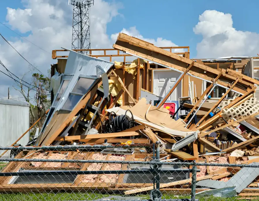 Wooden house debris scattered across a fenced site under clear skies after demolition.