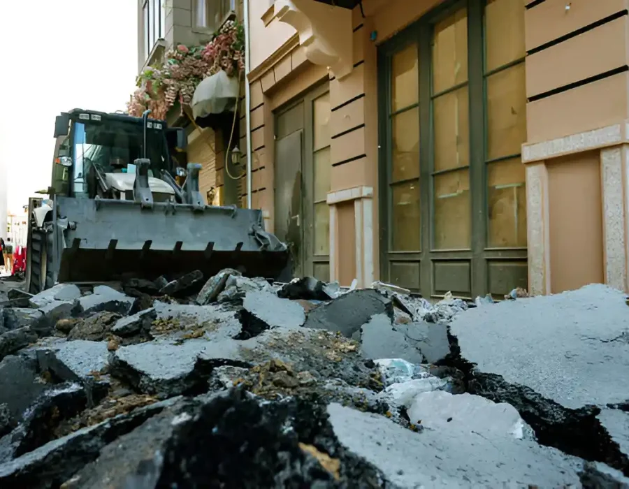 Bulldozer removing broken asphalt rubble from a street near buildings during cleanup operations.