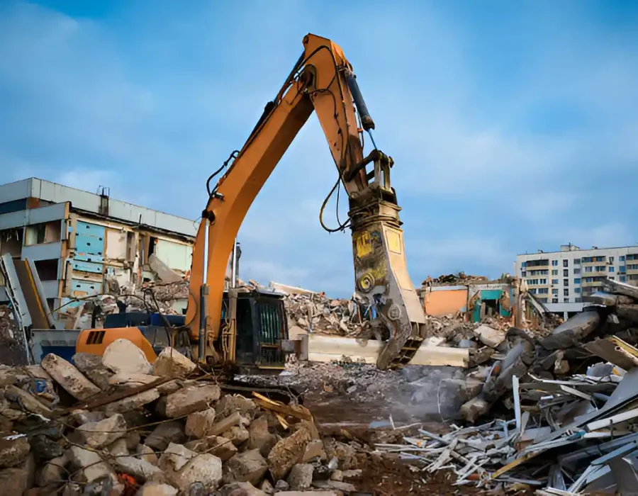 Yellow excavator clearing rubble at an urban demolition site with partially demolished buildings nearby.