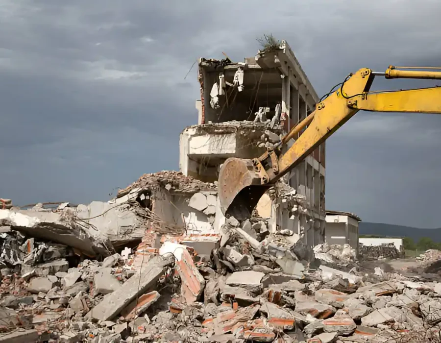 Yellow excavator tearing down a partially collapsed concrete building amid rubble and debris.
