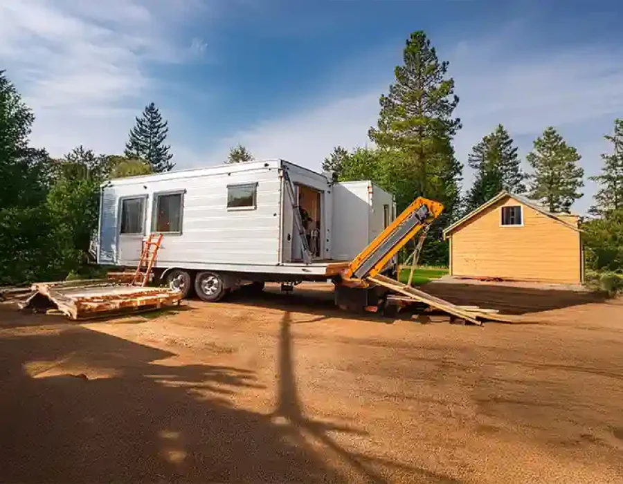 Construction crane positioned next to white mobile home with lumber debris on prepared forest lot