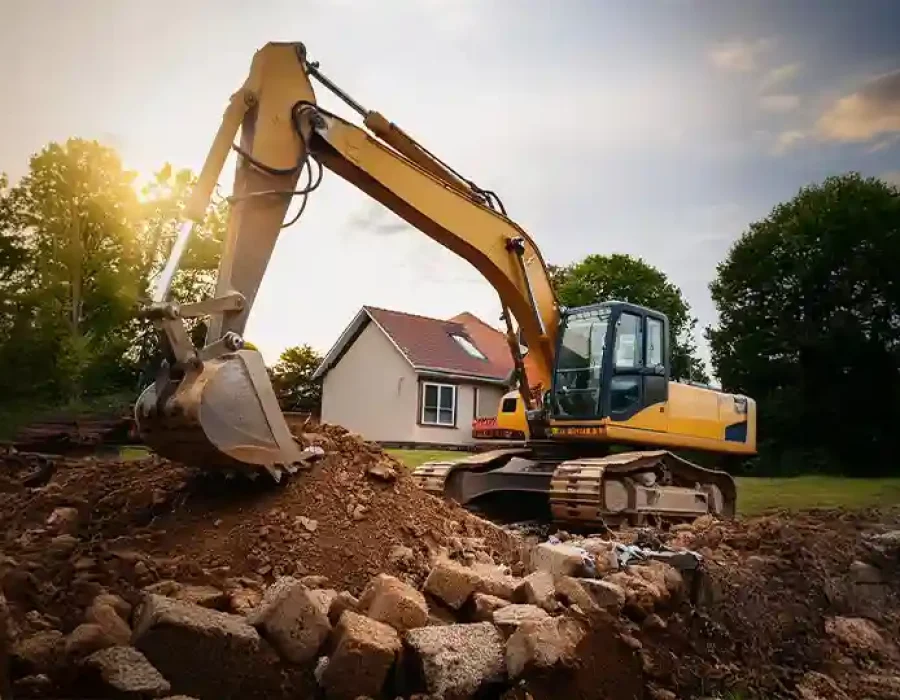 Yellow excavator removing rubble near residential house at a construction or demolition site.
