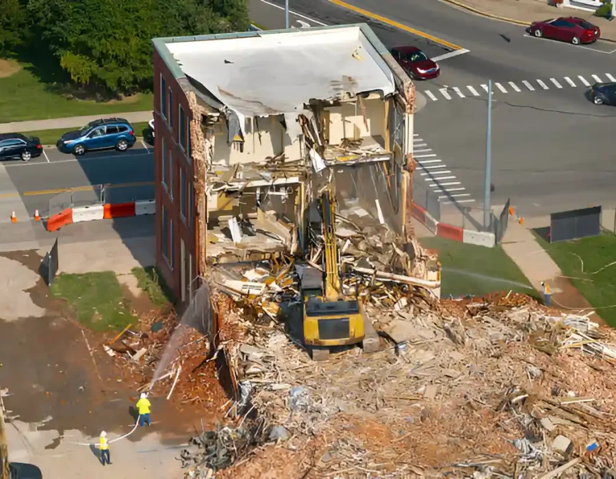 Excavator demolishing brick building with debris pile and workers managing commercial demolition site cleanup