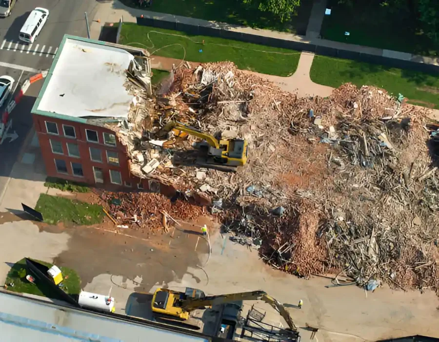 Aerial view of excavator demolishing brick building with extensive debris field and cleanup equipment on urban site