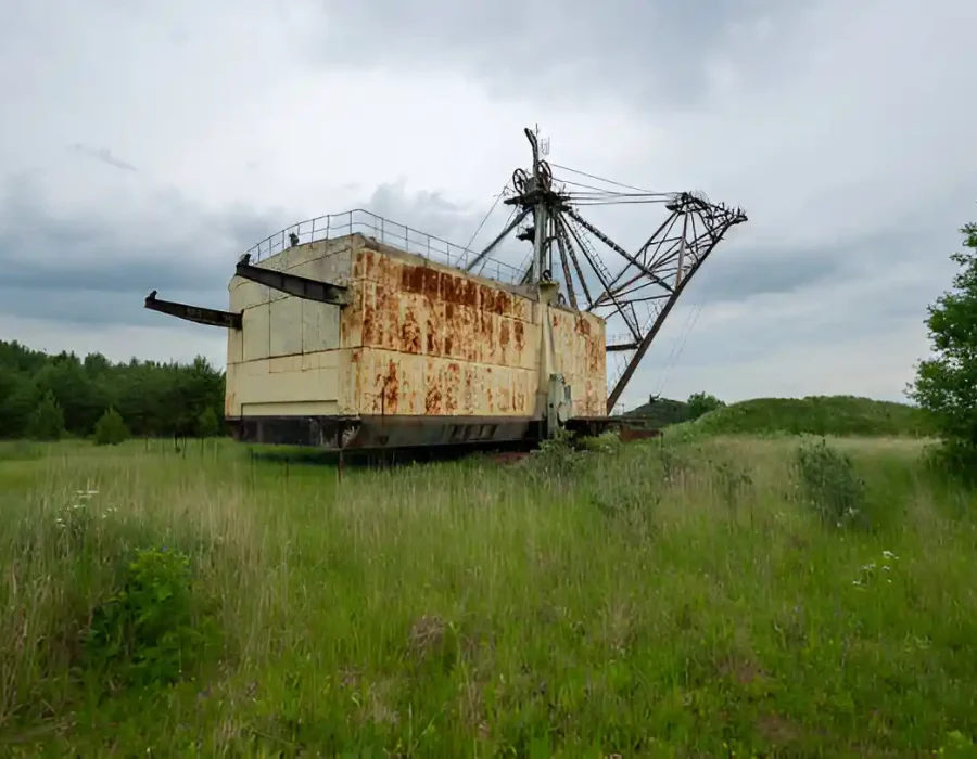 Rusty excavator abandoned in a grassy field under cloudy skies.
