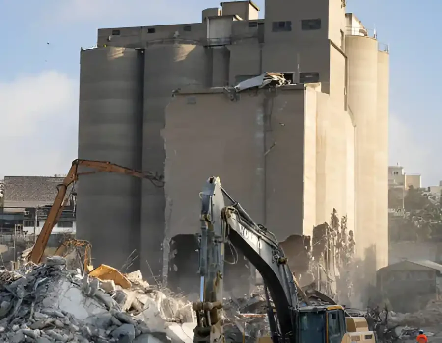Excavators demolishing a large industrial building with debris and workers in safety gear.