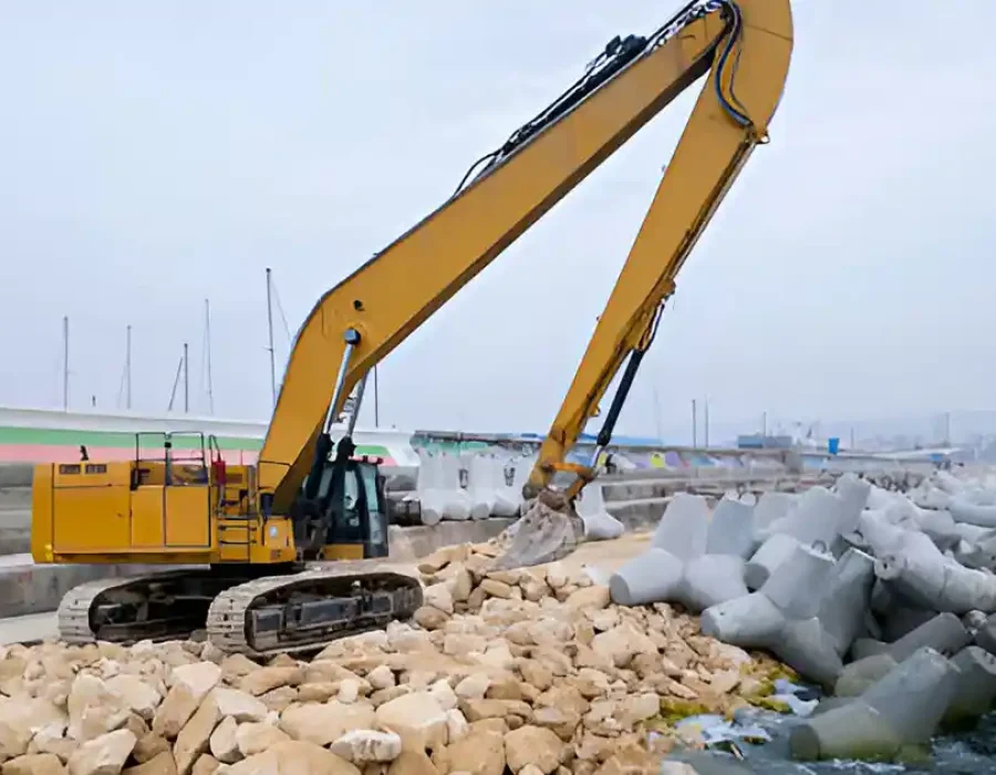 Excavator clearing rocks and debris at a coastal construction site.
