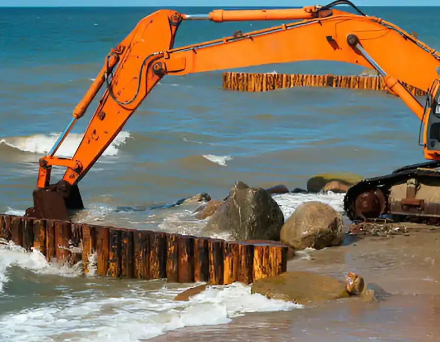 Orange excavator clearing wooden seawall, moving large rocks in coastal construction