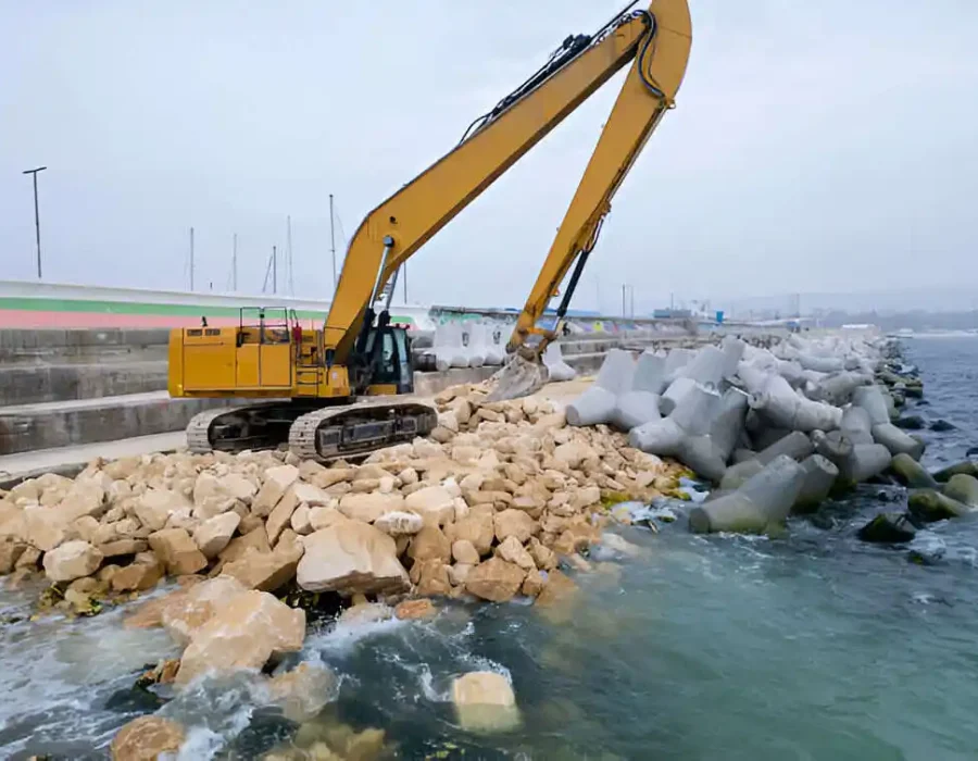 Yellow excavator moving rocky debris near concrete seawall, marine construction site