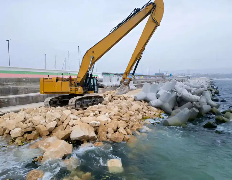 Yellow excavator positioning large rocks on a shoreline for coastal reinforcement and erosion control.