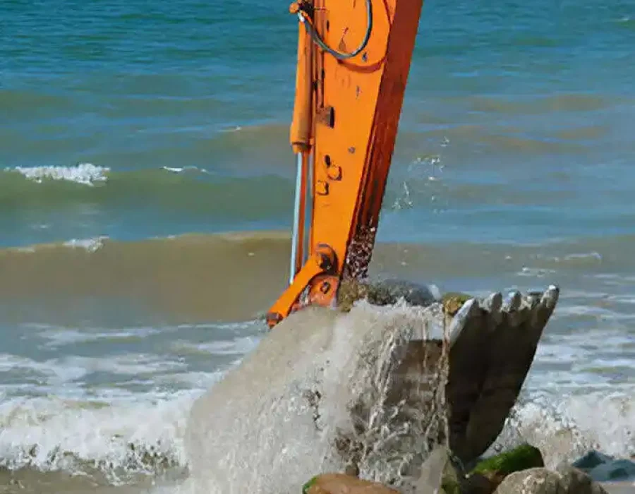 Orange excavator arm moving rocky debris in ocean shoreline wave splash